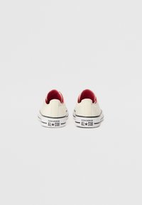 Béžové plátěné tenisky s červeným interiérem, černými pruhy na podrážce a logem "Converse All Star" na patě. Design s zaoblenou špičkou.