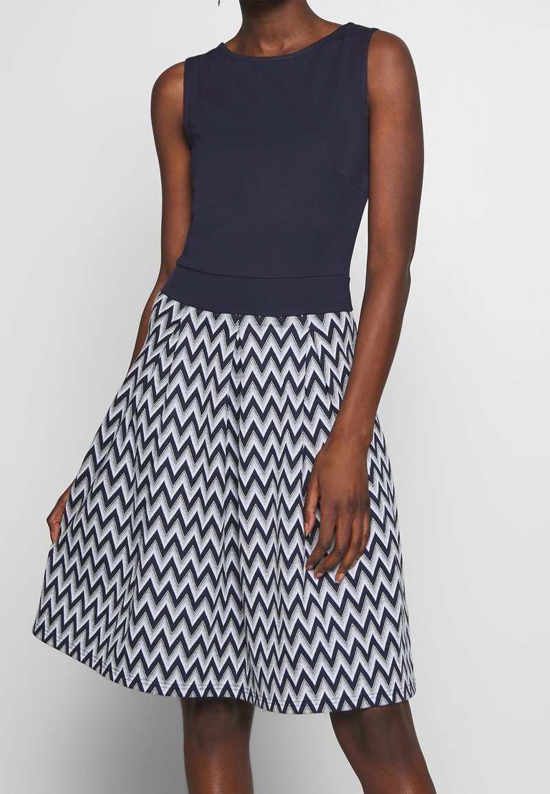 Femme portant une robe sans manches bleu marine avec une jupe à motif zigzag noir et blanc, debout devant un fond uni gris clair.
