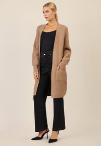 Cardigan lungo beige con texture a coste, apertura frontale e tasche laterali, abbinato a jeans neri a zampa con decorazioni.