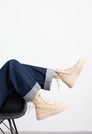 Zapatillas altas beige con suela gruesa y texturada, cordones en la parte frontal y un detalle de cremallera. Llevadas con unos jeans oscuros de mezclilla doblados.