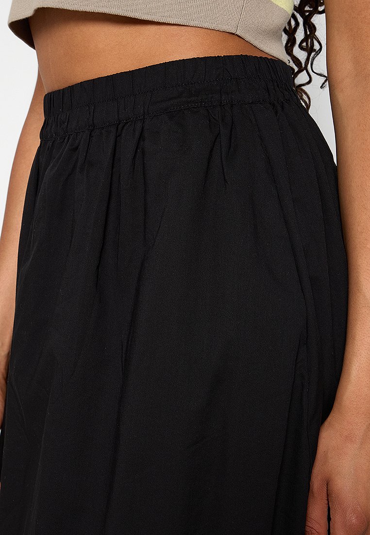 Czarna spódnica posiada elastyczny pasek w talii z marszczeniem. Wykonana z lekkiego materiału, ma gładką teksturę i swobodny krój.