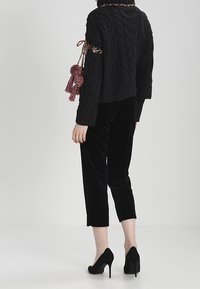 Maglione nero a trecce con frange decorative sulle maniche, abbinato a pantaloni neri in velluto taglio corto e scarpe con tacco nere.