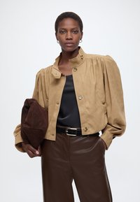 Γυναίκα με μπεζ σακάκι με κουμπιά και φουσκωτά μανίκια, μαύρο μεταξωτό τοπ, καφέ δερμάτινο παντελόνι, κρατώντας μια μεγάλη σκούρα καφέ σουέντ τσάντα χειρός.