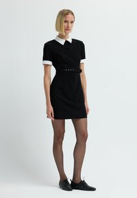 Donna in piedi che indossa un vestito nero a maniche corte con colletto e polsini bianchi, cintura nera, collant trasparenti e scarpe nere con lacci.