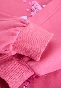 Primer plano de mangas de sudadera rosa con puños acanalados y lentejuelas rosas y moradas dispersas sobre fondo de tela.