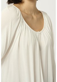 Femme portant un chemisier blanc texturé et ample avec des détails de tissu froncé et un petit bouton près de l'encolure, sur un fond uni.