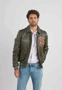 Veste aviateur en cuir vert foncé avec poignets côtelés, arborant un logo brodé sur la poitrine et des accents beige, portée avec une chemise claire et un jean.