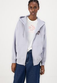 Sudadera gris con cremallera y una gran capucha, dos bolsillos delanteros y detalles bordados. Lleva una camiseta blanca debajo y está combinada con pantalones de mezclilla oscuros.