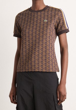 Personne portant une chemise marron à manches courtes avec un petit logo crocodile vert et des rayures violettes et jaunes sur les épaules.