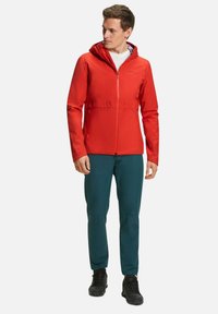 Giacca rossa con cappuccio, zip frontale, polsini elasticizzati e due tasche laterali, abbinata a pantaloni verde scuro e scarpe nere. Tessuto liscio e leggero.