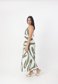 Abito maxi senza maniche bianco con motivo astratto verde. Tessuto morbido, forma a linea A e scollatura profonda sul retro. Un paio di sandali con tacco alto argento.