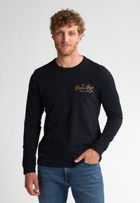 Zwarte langharige shirt gemaakt van katoen met een ronde hals en een gouden logo op de voorkant. Gepaard met blauwe jeans met een ontspannen pasvorm.