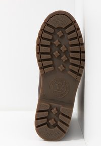 Semelle en caoutchouc marron d'une chaussure présentant des crampons profonds en forme de croix et un logo circulaire indiquant "Natural Life & Style Since 1989."
