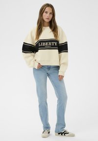 Sweatshirt crème avec des rayures noires, comportant les inscriptions "LIBERTÉ" et "UNIVERSITÉ". Un jean bleu clair et des baskets à rayures noires complètent la tenue.
