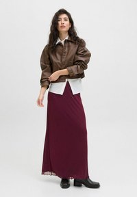 Chaqueta de cuero marrón corta con botones y bolsillos, superpuesta sobre una camisa blanca, combinada con una larga falda burdeos y botas negras hasta el tobillo.
