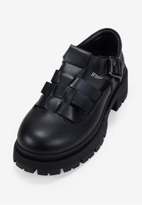 Zapato de cuero negro con diseño de correa trenzada, punta redonda, suela de goma gruesa y detalle de hebilla ajustable en el costado. Acabado texturizado.