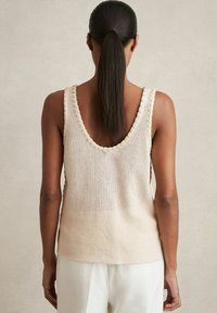 Beige gestrickter Tanktop mit weitem Ausschnitt, strukturiertem Netzmuster und Rüschendetails entlang der Kanten. Offenes Rückendesign sichtbar.