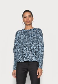 Blusa azul e preta com padrão, mangas longas, corpo franzido e decote redondo. Combinada com calças pretas. Tecido de textura suave.