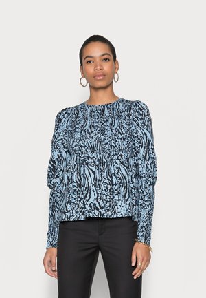 Blusa azul e preta com padrão, mangas longas, corpo franzido e decote redondo. Combinada com calças pretas. Tecido de textura suave.