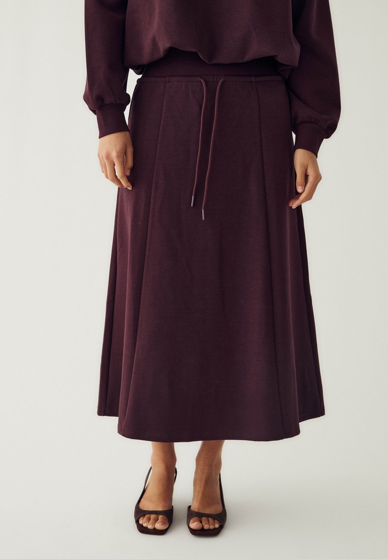 Femme portant une longue jupe violet foncé avec une taille à cordon et un haut à manches longues assorti, associée à des talons ouverts foncés.