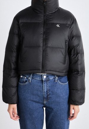 Zwarte korte donzen jas met hoge kraag en ritssluiting, gedragen met blauwe spijkerbroek. Klein "cK" logo op de linkerborst.