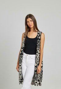 Schwarze und cremefarbene Häkelweste mit floralen Mustern, offenes Vorderdesign und spitzenähnliche Textur, kombiniert mit einem schwarzen Oberteil und weißen Hosen.