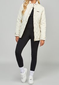 Quiltad beige jacka med ståkrage, tryckknappar och sidofickor, kombinerad med svarta leggings och vita sneakers.