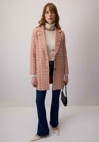 Kockovaný blejzer v oranžovej a smotanovej farbe, navrstvený na smotanovom roláku. Rozšírené denimové džínsy a špicaté biele topánky, v ruke drží čiernu tašku.