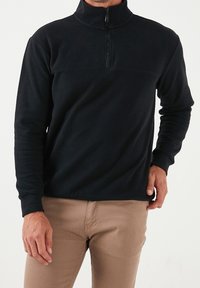 Pullover in pile nero con colletto a mezza zip, polsini a coste e texture liscia. Design semplice con una cucitura orizzontale sul petto.