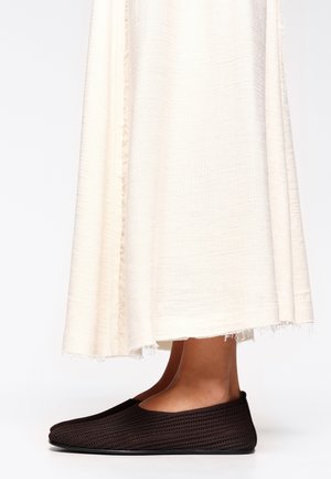 Jambes nues chaussées de mocassins en tricot marron foncé sous une longue jupe texturée couleur ivoire avec des bords effilochés.