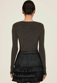 Naine, kellel on punutud kukk ja seljas pika särgiga sädelev top ning must kihiline fringe seelik, nähtud tagant ühetahelise tausta ees.