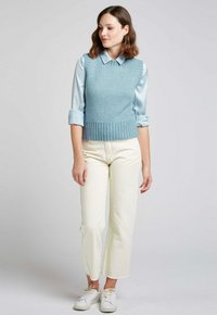Un pull sans manches en maille bleu clair avec un ourlet côtelé, porté sur une chemise à col bleu clair, associé à un pantalon blanc longueur cheville et des baskets blanches.