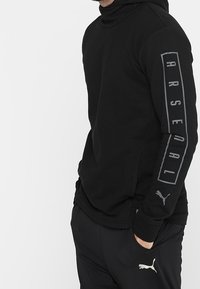 Sudadera negra con cuello alto, que presenta una impresión gris de "ARSENAL" en una manga y un pequeño logo de Puma en la otra manga. Tejido suave.
