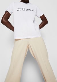 Camiseta blanca de algodón con el logotipo "Calvin Klein" en negro, combinada con pantalones de pierna ancha beige, textura suave y un diseño clásico de cuello redondo.