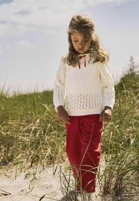 Vitt stickat tröja med öppet mönster och röd banddetalj, kombinerat med klara röda byxor, stående på ett gräsområde på en strand.