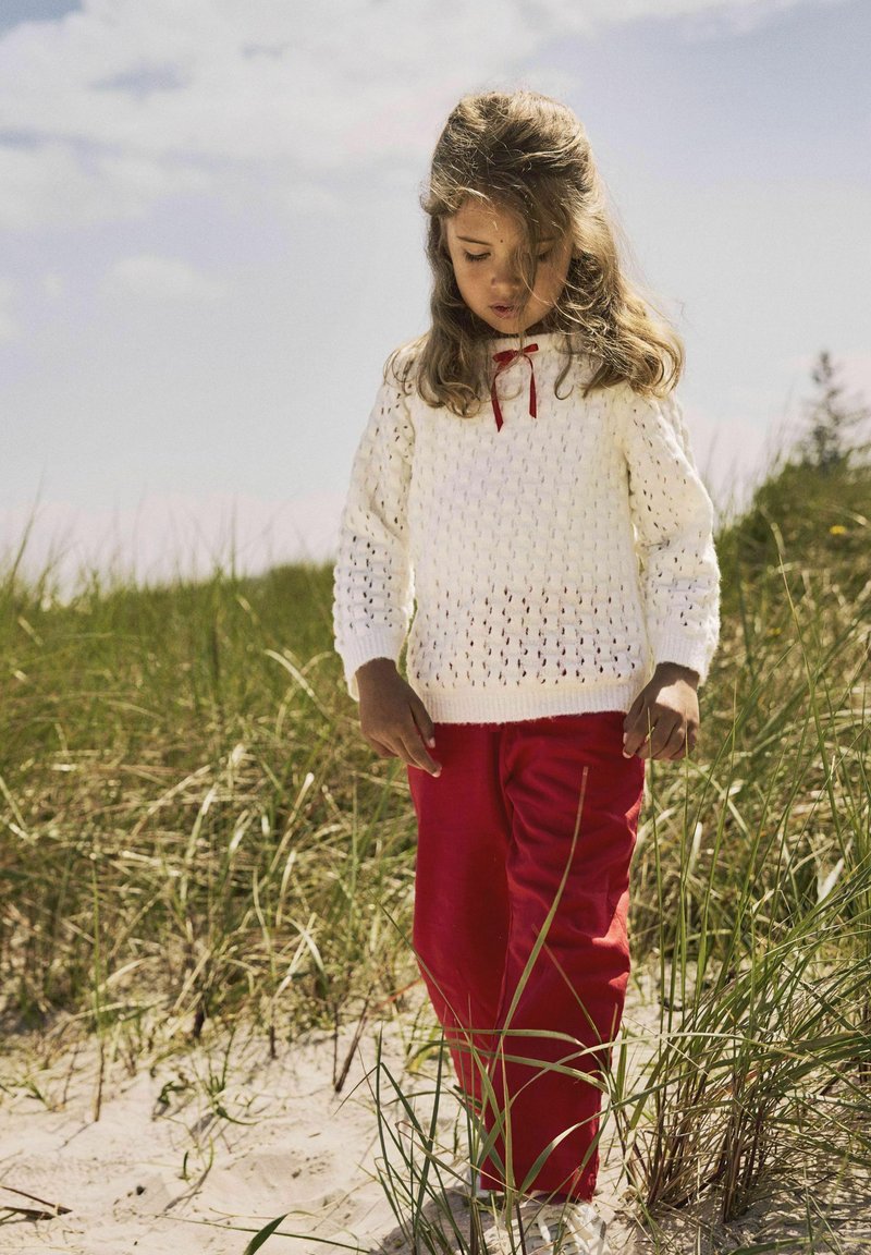Vitt stickat tröja med öppet mönster och röd banddetalj, kombinerat med klara röda byxor, stående på ett gräsområde på en strand.