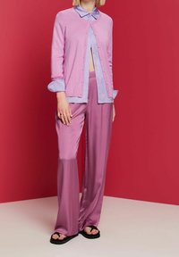 Růžový pletený cardigan přes světle modrou košili, doplněný o růžové saténové široké kalhoty a černé žabky na červeném pozadí.
