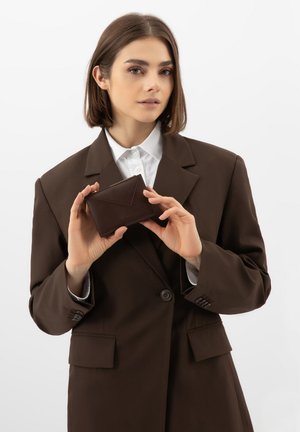 Frau in braunem Blazer, die mit beiden Händen eine kleine braune Lederbrieftasche vor einem schlichten weißen Hintergrund hält.