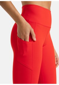 Leggings rouges taille haute en matière extensible, dotés d'une poche latérale, d'une texture lisse et de coutures plates pour le confort.