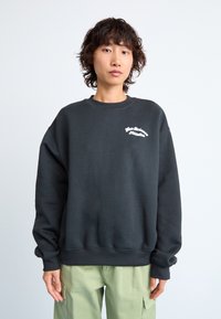 Svart oversized genser laget av myk stoff med en hvit tekstlogo på brystet. Ribbet mansjetter og klassisk rund halsdesign.