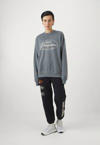 Šedá mikina s bílým logem, doplněná tmavě modrými tepláky s různými barevnými nášivkami a bílým nápisem. Bílé tenisky.