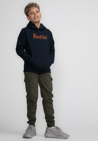 Marineblauwe hoodie met oranje "Petrol Ind" logo, gecombineerd met olijfgroene cargobroek met zakken, en grijze sportschoenen.