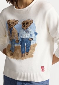 Bílý pletený svetr s výšivkou dvou medvědů v běžném oblečení, s modrými akcenty a červeným logem na lemu.