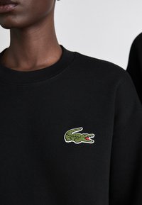 Crni dukserica s logotipom zelenog krokodila i crvenim detaljima, izvezenim na lijevoj strani prsnog područja, s rebrastim ovratnikom.