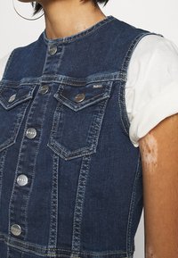 Sukienkaogrodniczka z denimu charakteryzuje się ciemnoniebieskim kolorem, strukturalnym fasonem, srebrnymi guzikami, podwójnymi kieszeniami na piersi oraz okrągłym dekoltem, pod którym widoczna jest biała koszula.