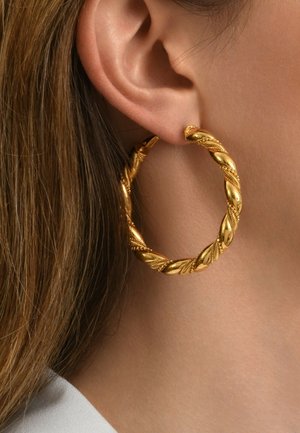 Boucles d'oreilles en or en forme de créole avec un design torsadé, présentant une surface texturée et de petites nervures, suspendues à une oreille percée.