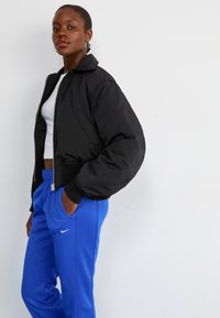 Black bomber jacket with ribbed cuffs and collar, paired with blue athletic pants featuring a white Nike logo.