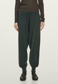 Persoon in losse donkergrijze joggingsbroek, bruin langmouwtop en zwarte instap schoenen, staande tegen een effen lichte achtergrond.