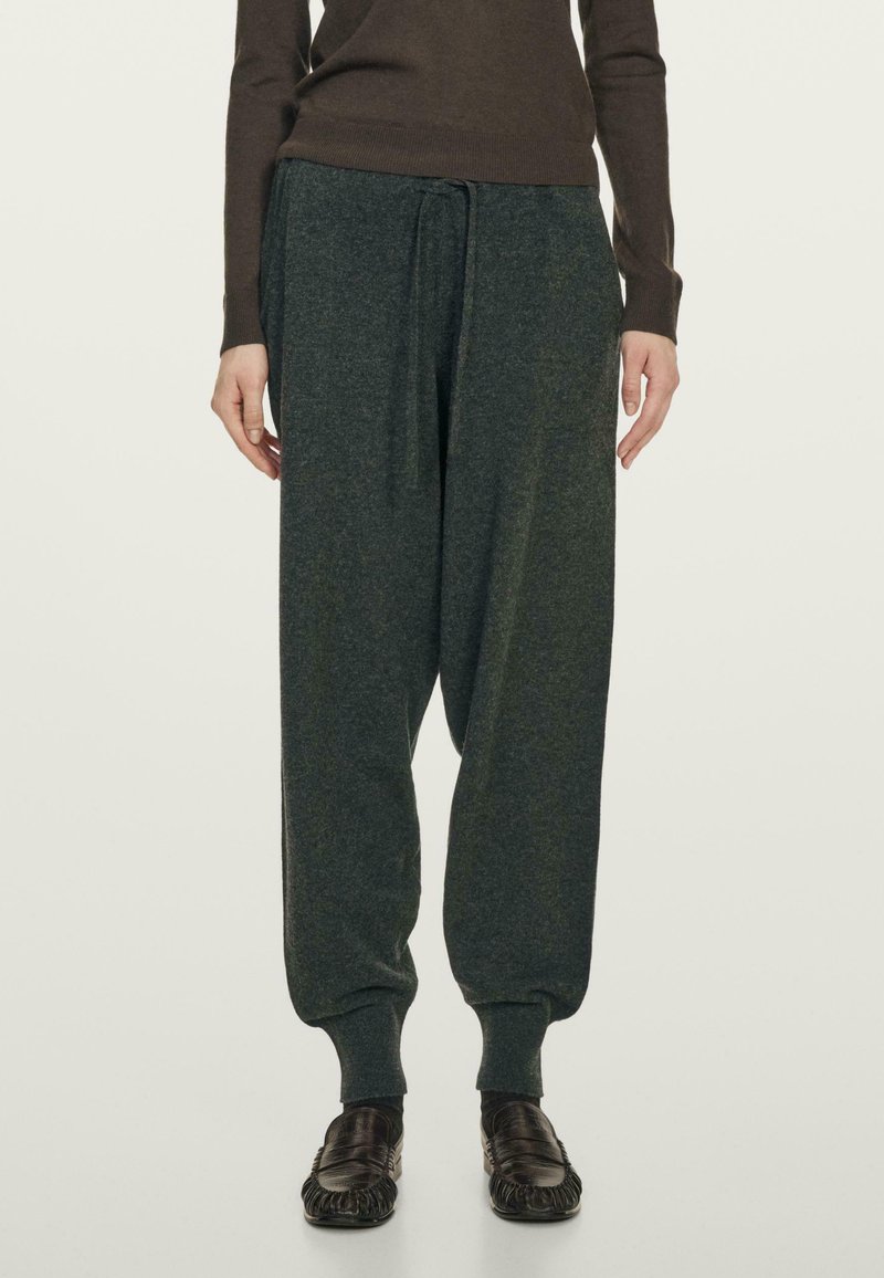 Persoon in losse donkergrijze joggingsbroek, bruin langmouwtop en zwarte instap schoenen, staande tegen een effen lichte achtergrond.