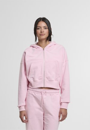 Felpa corta rosa con zip, tasche frontali e coulisse. Realizzata in tessuto morbido con una consistenza liscia. Abbinata a pantaloni coordinati.